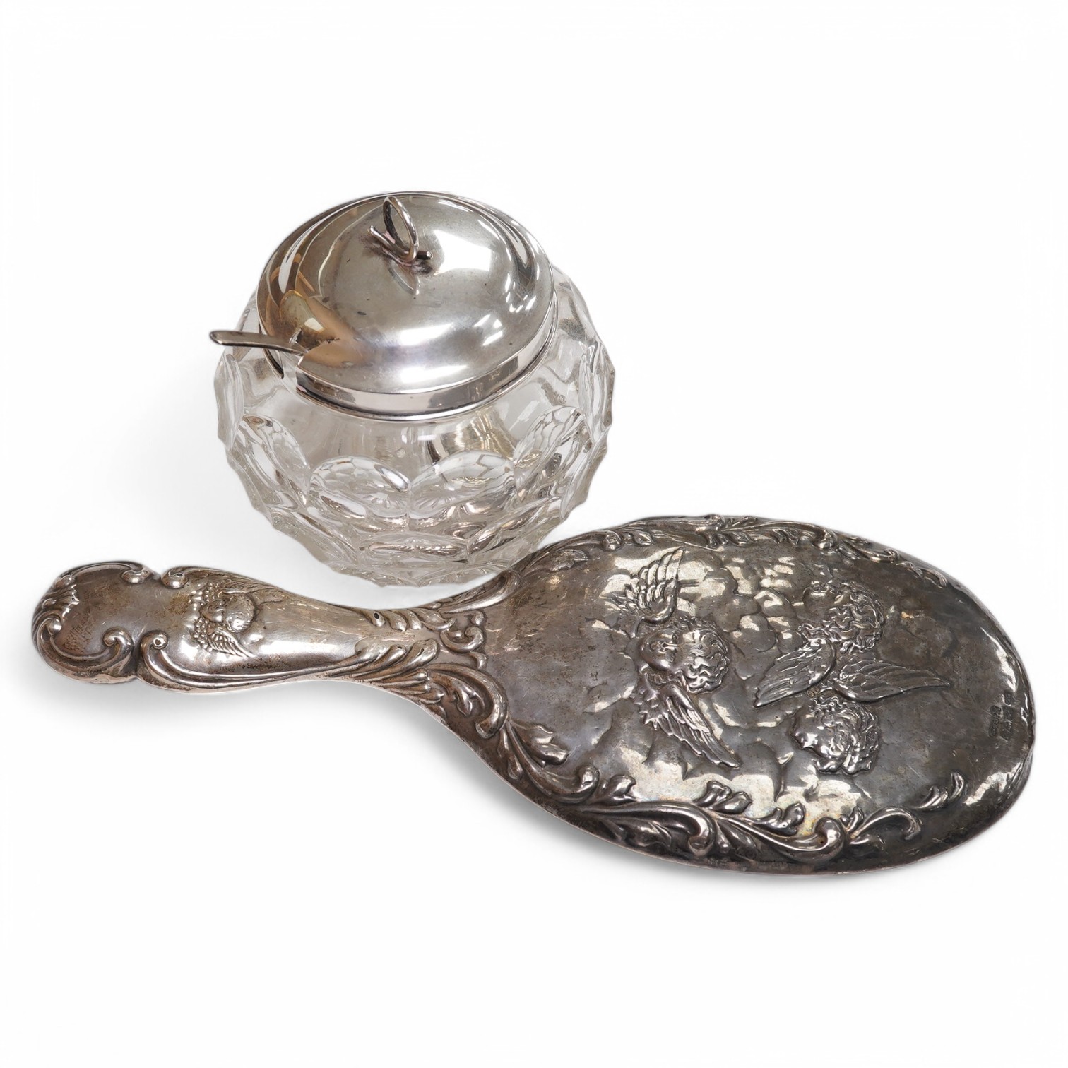 An Edwardian silver mounted hand mirror with Reynold's Angels decoration, William Neale Ltd, Chester, 1902, 26.3cm, together with a later silver mounted cut glass preserve jar and spoon. Condition - poor to fair